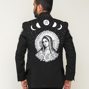 May include: Black blazer with a detailed white illustration on the back. The design features a religious figure surrounded by a halo, roses, moons, and stars. The blazer has a classic tailored fit.
