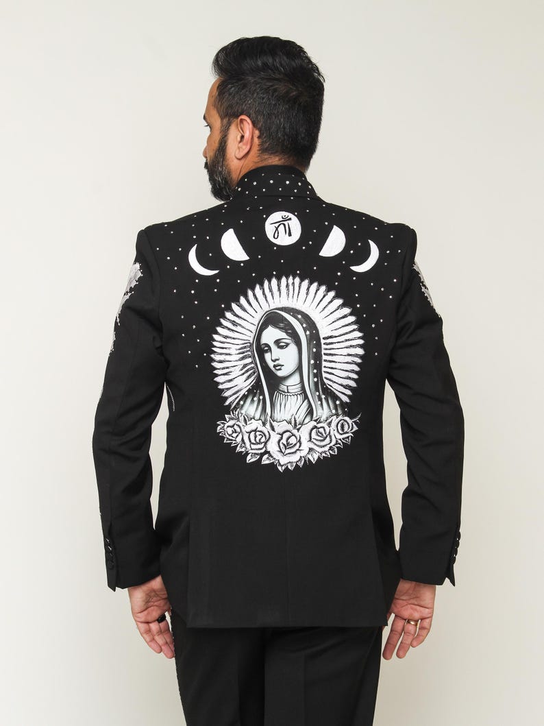 May include: Black blazer with a detailed white illustration on the back. The design features a religious figure surrounded by a halo, roses, moons, and stars. The blazer has a classic tailored fit.