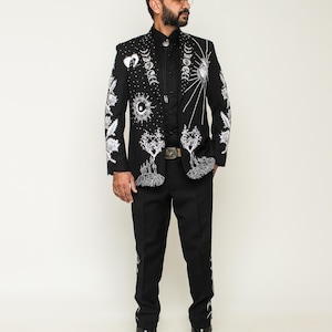 May include: A black suit with white celestial embroidery, including moons, suns, and trees. The suit features a matching jacket and pants, with detailed designs on the sleeves and legs. The outfit is completed with a black shirt and boots.