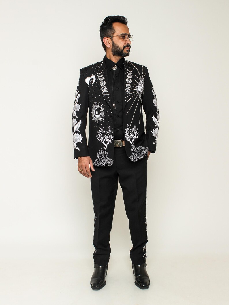 May include: A black suit with white celestial embroidery, including moons, suns, and trees. The suit features a matching jacket and pants, with detailed designs on the sleeves and legs. The outfit is completed with a black shirt and boots.