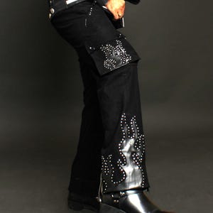 May include: Black denim pants with a flame design on the side. The design is made of black leather and has silver studs. The pants have a wide leg and are worn with black boots.