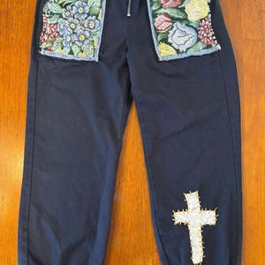 May include: Navy blue pants with embroidered floral pocket details and a white cross applique on the leg. The pants have a zipper fly and elastic cuffs. The pockets feature colorful floral embroidery.