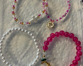 Könnte beinhalten: Vier Armbänder mit Perlen in verschiedenen Stilen. Ein Armband hat weiße Perlen, ein anderes rosa und transparente Perlen mit einem Kreuzanhänger, und ein drittes hat einen Kreuzanhänger. Das vierte Armband besteht aus großen rosa Perlen.