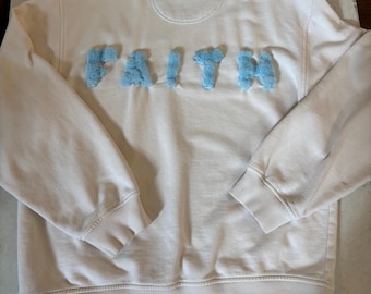 May include: Cream-colored sweatshirt with the word "FAITH" in light blue, fluffy, textured letters across the chest. The sweatshirt has a crew neck, long sleeves, and ribbed cuffs and hem.