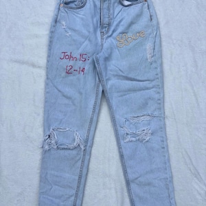 May include: Pale blue denim jeans with ripped details. The jeans have the word "Love" in gold sequins and the text "John 15: 12-14" in red. The jeans have a high waist and a straight leg.
