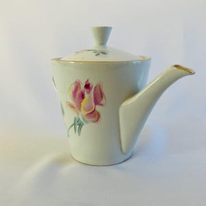 May include: Cream-colored ceramic teapot with a lid and a spout. The teapot is decorated with a pink and yellow rose design. The spout and rim are trimmed with gold. The teapot is on a white surface.