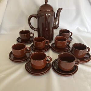 May include: A glossy dark brown ceramic coffee set consisting of a tall lidded coffee pot with a curved handle and spout, accompanied by seven matching cups and saucers. Each cup features a rounded body and a small side handle, resting on a plate.