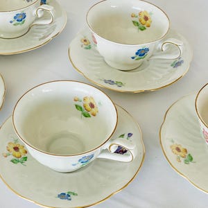 Può includere: Un set di tazze e piattini in porcellana bianca. Ogni tazza e piattino è decorato con un motivo floreale giallo, blu e rosa, con bordi dorati. Le tazze hanno manici curvi e poggiano su piattini con bordi smerlati.