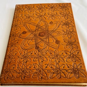 May include: A closed, hardbound book with a brown leather cover. The cover features an embossed design of atoms and atomic structures. The book's edges are also brown, and the overall aesthetic is vintage.
