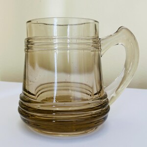 Peut inclure: Une chope en verre ambré d'époque avec une anse. La chope a un corps cylindrique avec des rainures horizontales près de la base et du sommet. La poignée est incurvée et fixée sur le côté. Le verre est transparent.