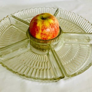 Op de afbeelding: Een serveerschaal van helder glas met een rond middengedeelte en vijf uitstralende secties. Het glas is voorzien van een gestructureerd, geribbeld patroon langs de randen en de bodem. Een rode en gele appel ligt in de middelste kom.