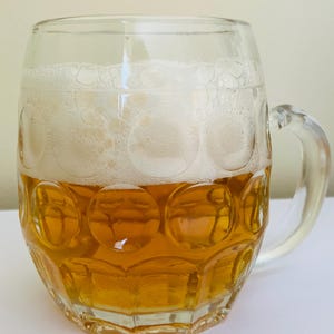 Op de afbeelding: Een heldere bierpul gevuld met goudkleurig bier en bedekt met een dikke laag wit schuim. De pul heeft een handvat en een decoratief patroon van cirkels rond de basis.