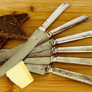 May include: A collection of silver butter knives arranged on a wooden cutting board, alongside two slices of dark bread and a square of yellow butter. The knives have a classic design with a slightly curved blade and handle.