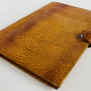 May include: A rectangular, textured, brown leather wallet with a reptile-like pattern. The wallet has a flap closure with a small, round button. The leather has a gradient of colors, from light to dark brown.