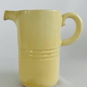 Könnte beinhalten: Eine hellgelbe Keramikkrug mit rundem Körper und gebogenem Griff. Der Krug hat einen Ausguss zum Ausgießen und zwei horizontale Rillen in der Mitte. Das Design ist schlicht und klassisch, geeignet zum Servieren von Getränken.