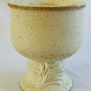 Könnte beinhalten: Cremefarbene Keramik-Sockelschale mit strukturierter Oberfläche. Die Schale hat eine breite, abgerundete Oberseite mit braunem Rand und steht auf einem Sockel mit Blattmuster. Ein dekoratives Stück für das Zuhause.