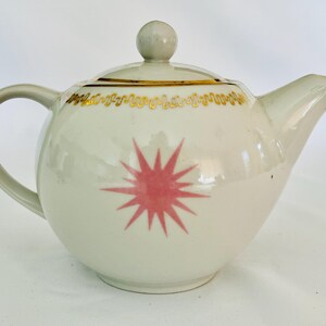 May include: A white ceramic teapot with a pink starburst design on the front. The teapot has a curved handle, a spout, and a lid with a small knob. Gold trim decorates the rim of the lid. A charming addition to any tea set.