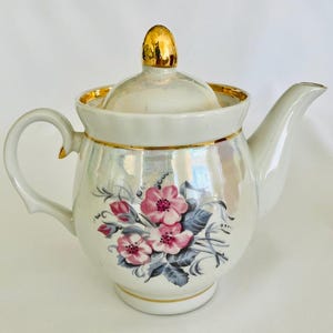 Può includere: Teiera in ceramica bianca con coperchio e finiture dorate. La teiera presenta un motivo floreale con fiori rosa e foglie grigie. Il manico e il beccuccio sono curvi e la teiera ha una finitura perlata.