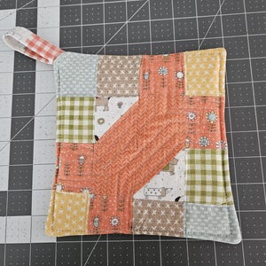 May include: A square, quilted pot holder with a hanging loop. The patchwork design features dog illustrations, floral motifs, and gingham patterns. The colour palette includes orange, yellow, green, and brown. The edges are bound with yellow fabric.