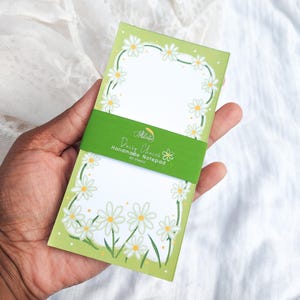 Pode incluir: Um bloco de notas verde e branco com um design de corrente de margaridas. O bloco de notas tem uma faixa verde com o texto "Daisy Chains Handmade Notepad 40 sheets". O bloco de notas é segurado em uma mão.