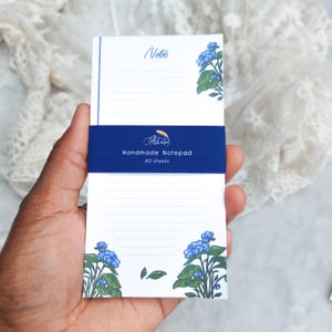 Pode incluir: Um bloco de notas artesanal com uma faixa azul e detalhes florais. O bloco de notas apresenta páginas pautadas e as palavras "Notes" e "Handmade Notepad" com o número de folhas impresso na frente. O design inclui flores azuis e folhas verdes.