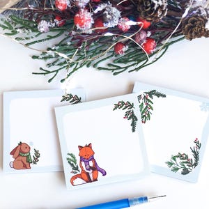 Può includere: Tre post-it quadrati azzurri con illustrazioni di un coniglietto, una volpe e un fiocco di neve. Il coniglietto e la volpe indossano sciarpe. Ogni nota ha un rametto verde e rosso di foglie e bacche. Una penna blu è in primo piano.