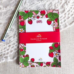 Può includere: Un blocco note rettangolare con un bordo di piante di fragole con bacche rosse, foglie verdi e fiori bianchi su sfondo rosa. Una fascia di carta rossa avvolge il centro con il testo Handmade Notepad 40 sheets. Una penna a righe verdi e bianche.