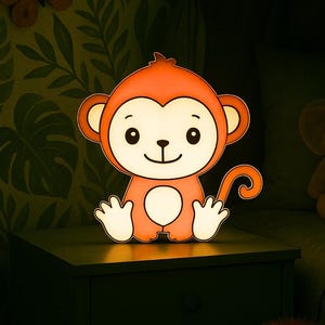 Puede incluir: Una luz de noche con forma de mono naranja brillante. El mono de dibujos animados tiene una cara sonriente, orejas grandes y una cola rizada. Está sobre una superficie oscura en una habitación con poca luz.