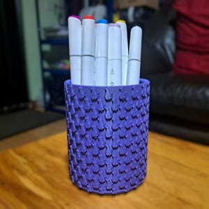 Pode incluir: Um porta-lápis cilíndrico roxo, texturizado, com uma variedade de marcadores coloridos dentro.