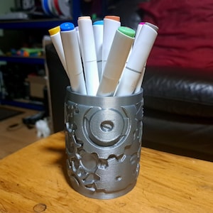 May include: A silver-colored pen holder with a gear design, filled with various colored markers. The markers have white barrels and colorful caps. The pen holder is cylindrical and sits on a wooden surface.