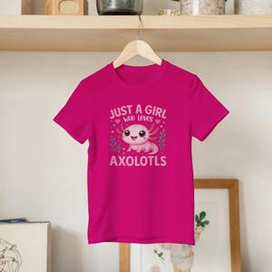 Könnte beinhalten: Ein leuchtend magentafarbenes Kurzarm-T-Shirt mit einer zentralen Grafik eines lächelnden rosa Axolotls, umgeben von kleinen Herzen und Blattzweigen. Der in Weiß gedruckte Text lautet "JUST A GIRL WHO LOVES AXOLOTLS" über und unter der Abbildung.