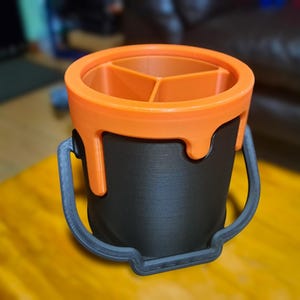 Puede incluir: Un organizador de escritorio impreso en 3D con forma de cubo de pintura. El cubo es negro con una parte superior naranja que tiene tres compartimentos. El asa y la base son grises. La parte superior naranja tiene un diseño de pintura goteando.