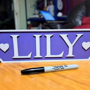 Op de afbeelding: Een paars houten bord met witte letters en witte hartjes aan beide kanten. Het bord heeft de tekst "LILY".
