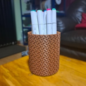 Puede incluir: Un porta lápices cilíndrico marrón texturizado con una variedad de marcadores de colores.