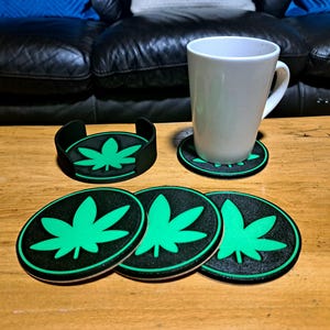 Könnte beinhalten: Ein Set aus schwarzen und grünen Cannabisblatt-Untersetzern und einem passenden Halter. Die runden Untersetzer haben einen schwarzen Hintergrund und ein grünes Blattdesign. Eine weiße Tasse steht auf einem der Untersetzer.