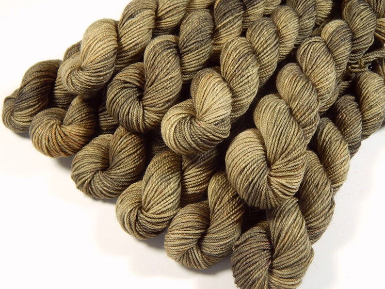 May include: A collection of multiple skeins of yarn twisted into bundles. The yarn features a variegated color palette consisting of earthy tones, including shades of dark brown, olive green, and light tan, creating a textured appearance.