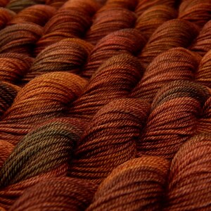 K&ouml;nnte beinhalten: Eine Ansammlung von gedrehten Garnstr&auml;ngen, die in einem sich wiederholenden Muster angeordnet sind. Das Garn weist eine satte Farbpalette aus tiefem gebranntem Orange, Kupfer und Dunkelbraun mit subtilen olivgr&uuml;nen Akzenten in den texturierten Fasern auf.