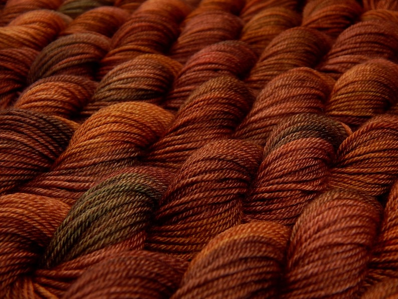 K&ouml;nnte beinhalten: Eine Ansammlung von gedrehten Garnstr&auml;ngen, die in einem sich wiederholenden Muster angeordnet sind. Das Garn weist eine satte Farbpalette aus tiefem gebranntem Orange, Kupfer und Dunkelbraun mit subtilen olivgr&uuml;nen Akzenten in den texturierten Fasern auf.