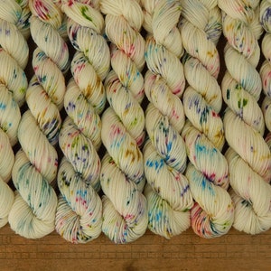 May include: A collection of off-white yarn skeins arranged in a row, each featuring a speckled pattern of blue, pink, green, and yellow dots. A wooden ruler is visible at the bottom, showing measurements in inches from 13 to 22 inches.