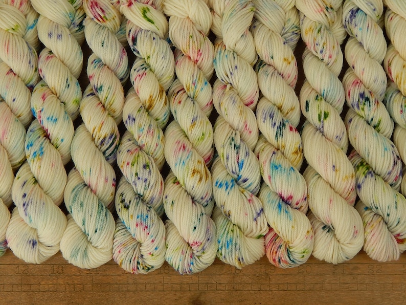 May include: A collection of off-white yarn skeins arranged in a row, each featuring a speckled pattern of blue, pink, green, and yellow dots. A wooden ruler is visible at the bottom, showing measurements in inches from 13 to 22 inches.