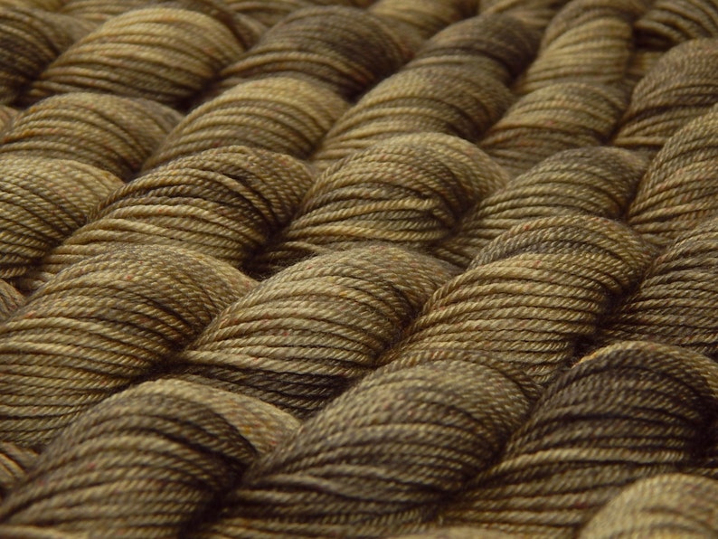 May include: Multiple skeins of yarn arranged in a repeating pattern. Each skein features a variegated color palette of dark brown, tan, and muted olive green tones, showcasing the twisted fiber texture and the individual strands of the yarn.