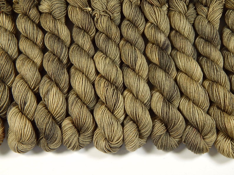 May include: A collection of multiple skeins of yarn arranged in a row. Each skein features a variegated color pattern with shades of olive green, brown, and tan, displaying a twisted, multi-ply texture throughout the length of the fiber.