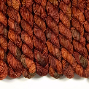K&ouml;nnte beinhalten: Eine Ansammlung mehrerer gedrehter Garnstr&auml;nge, die in einer Reihe angeordnet sind. Das Garn weist eine nuancierte Farbpalette aus tiefem gebranntem Orange, Kupfer und Rostt&ouml;nen auf, mit subtilen Akzenten in Dunkelbraun und gedecktem Olivgr&uuml;n.
