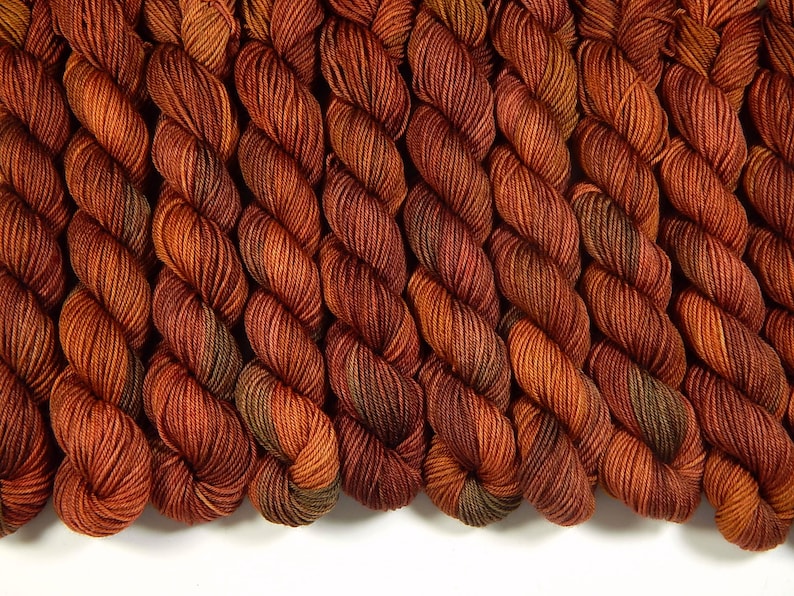 K&ouml;nnte beinhalten: Eine Ansammlung mehrerer gedrehter Garnstr&auml;nge, die in einer Reihe angeordnet sind. Das Garn weist eine nuancierte Farbpalette aus tiefem gebranntem Orange, Kupfer und Rostt&ouml;nen auf, mit subtilen Akzenten in Dunkelbraun und gedecktem Olivgr&uuml;n.