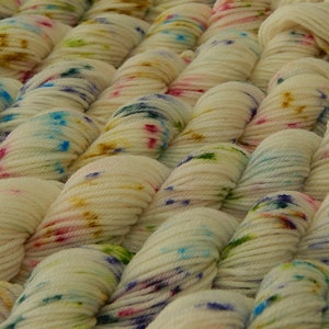 May include: A collection of off-white yarn skeins arranged in parallel rows. Each skein features a speckled pattern with small, scattered dots of blue, pink, green, and golden-brown dye, creating a textured appearance across the entire surface.
