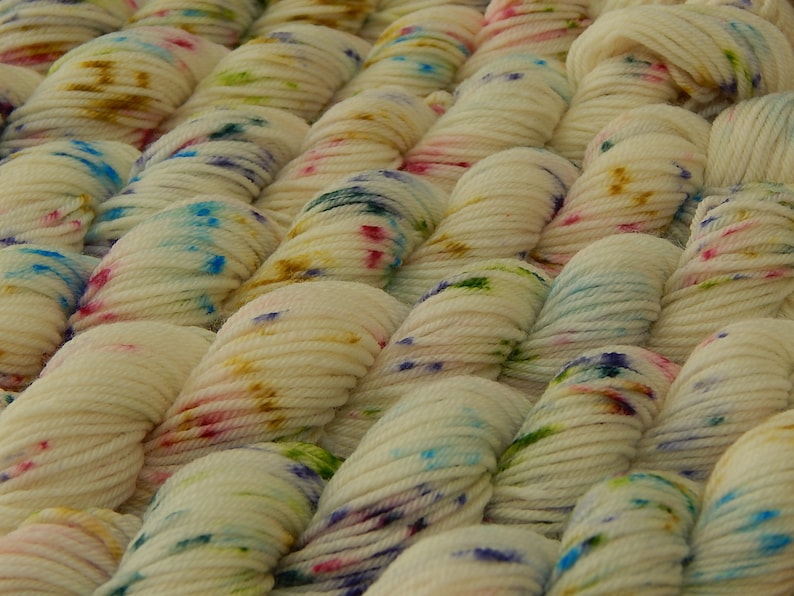 May include: A collection of off-white yarn skeins arranged in parallel rows. Each skein features a speckled pattern with small, scattered dots of blue, pink, green, and golden-brown dye, creating a textured appearance across the entire surface.