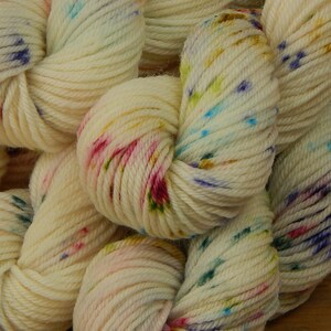 May include: A collection of off-white yarn skeins featuring a speckled pattern with small dots of blue, pink, yellow, and green. The yarn is twisted into soft, rounded bundles, showcasing a multi-colored, vibrant, and textured appearance throughout.