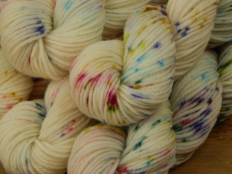 May include: A collection of off-white yarn skeins featuring a speckled pattern with small dots of blue, pink, yellow, and green. The yarn is twisted into soft, rounded bundles, showcasing a multi-colored, vibrant, and textured appearance throughout.