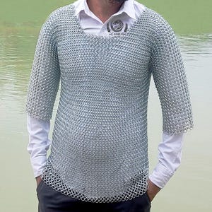 Puede incluir: Una camisa de cota de malla plateada con mangas cortas, usada sobre una camisa blanca con cuello. La cota de malla está hecha de pequeños anillos de metal entrelazados. La camisa blanca tiene un logotipo en el pecho. El fondo es una masa de agua.