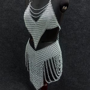 May include: A silver chainmail halter top and skirt set displayed on a black mannequin. The top features a plunging neckline and the skirt has a fringed hemline. The chainmail is made of small, interlocking metal rings.
